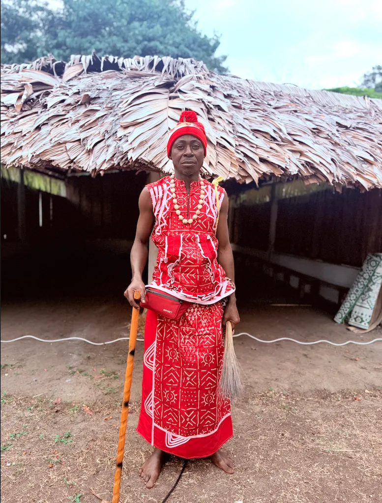 Jérôme Mouele Niangou devant le temple, en tenue rituelle rouge, bonnet et collier de graines, canne et chasse-mouches en main